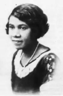 An African-American woman with wavy hair cut in a bob. She is wearing a necklace and a lace-collared top with a cut-out detail on the shoulder.