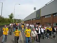 A group of people wearing predominantly yellow football shirts, walking along a road beside an association football stadium