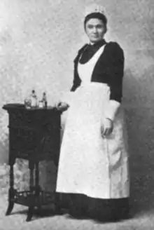A young woman with dark hair and brown skin, wearing a nurse's uniform, standing next to a small table with bottles on it