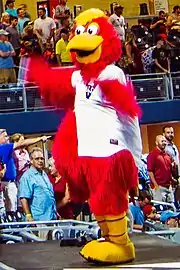 A person wearing a red anthropomorphized rooster costume dressed in a white baseball jersey with a blue "V" on the right chest dances on a baseball dugout.