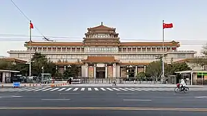An ornate building front stretching the width of the image, slightly tilted, against a uniformly blue sky. Its front has a projecting pavilion in the Chinese style, echoed by a similar pagoda-style top on the roof above it.