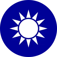 A blue circular emblem on which sits a white sun composed of a circle surrounded by 12 rays.