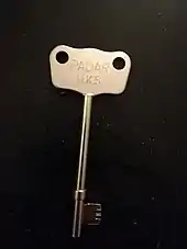  A large, silver-coloured key with a wide head is depicted on a dark-coloured background.On the head of the key, the text RADAR N.S.K is inscribed.