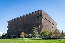 A three tiered golden colored building with no visible windows and a green lawn in front.