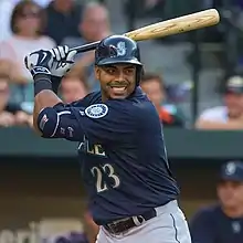 A man wearing a navy blue baseball jersey and batting helmet stands with his bat held back, awaiting a pitch.