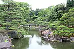 A pond with stones along the sides located in a garden with pine and other trees.