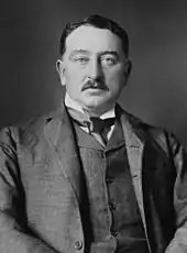 A moustachioed man in a dark three-piece suit