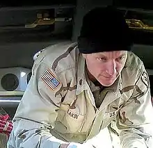 A caucasoid man in desert fatigues and a black knit cap is standing, bent-over towards the camera, and looking off center at an unpictured patient.