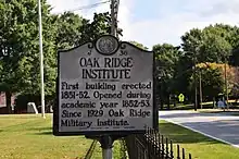 A small sign near a fence containing text. The text reads: OAK RIDGE INSTITUTE First building erected 1851-1852 Opening during academic year 1852-1853. Since 1929, Oak Ridge Military Institute