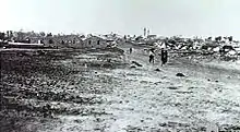 In the distance a group of people walk down a dirt road away from the village. The foreground shows vacant ground.
