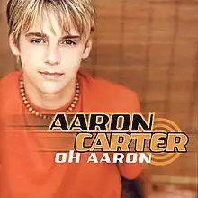 A young male adult with blonde hair is wearing an orange t-shirt and a bead necklace. The artist's name is colored in black and orange, and the album title is colored in white.