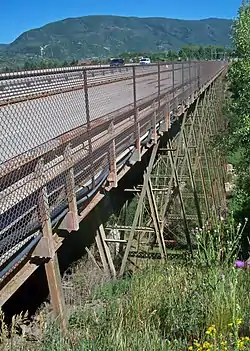 An old metal bridge across a grassy, wooded gorge, seen from its right. Diagonal supports go down below the bridge. There is a wooded mountain in the background.