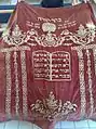 Parochet in red hue decorated with embroidery of The Ten Commandments. Memorial for Reuven ben Zmra, died on 1924.
