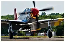 A single nosed-propeller aircraft is on the ground on its wheels with the propeller in motion.  The aircraft is viewed from the front, but the red nose faces slightly to the right.  The aircraft has black propellers.  Parts of the wings and propellers are cropped from view.