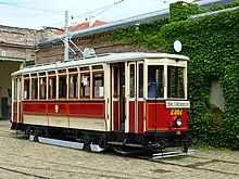 An antique ivory and white single streetcar in front of a brick building. The car has the crest of Vienna on its side, and the title "Erdberg Straßenbahnmuseum" on its front. The tram is empty except for a driver.