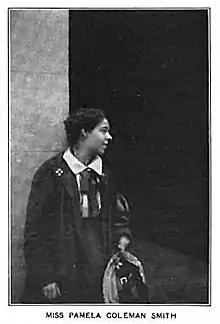 Pamela Colman Smith standing, dressed in masculine type clothes, holding a hat