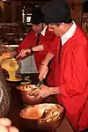 Two chefs in red smocks and black berets, one stirring eggs in a copper bowl and the other pouring batter from a bowl into a pan