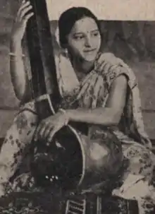 A young South Asian woman wearing a light-colored sari, seated, arms around a large stringed instrument