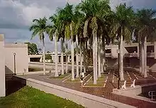 A grid of palm trees arranged in a tiled courtyard stands to the right of a dormitory building.