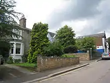Photograph of several houses, partly hidden by trees, behind a garden wall on an upward-sloping suburban road