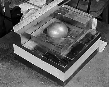 A square base of metal blocks, with a smaller square of metal on the top in the center, a metal ball (the "core") contained in its center. A ruler along one side of the base shows it is roughly 10.5 inches (270 mm) square.