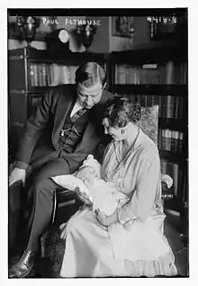 A white man in a suit and a white woman in a dress; the woman is holding a baby, presumably their daughter; both adults are looking at the baby. They are posed in front of a bookcase.