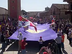 People at a celebration holding a huge purple flag. Others wave different flags, such as the Cross of Burgundy or the modern flag of Castile and León.