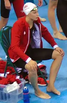 Penny Oleksiak in a swimsuit and jacket, seated on a bench by the Olympic pool.