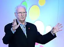 A photograph of Pete Waterman, an elderly Caucasian man with white hair, wearing a suit, sitting with his arms folded