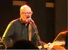 Holsapple singing into a microphone while playing guitar