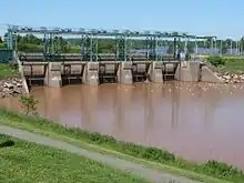 A concrete and steel structure crosses the river. The water is brown on the near side of the structure, and blue on the far side