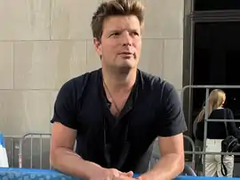 A dark-haired man wearing a black shirt rest his arms in a fence