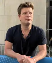 A dark-haired man wearing a black shirt rest his arms in a fence