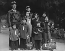 Dorothea Lange's photo of the Mochida family waiting for the bus in Hayward (1942). Hiroko and Miyuki are in the front row, at left; Miyuki is holding a sandwich. The children are wearing tags with the family number to help keep them together.