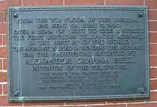 An image of darkened brass historical plaque with a streak of green corrosion running down it, mounted on the exterior side of a brick building.