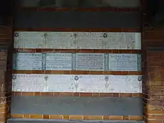Memorial tablets arranged in three rows on a wall. Those in the centre are in the green and white Arts and Crafts Movement style, while those above and below are in the more recent design. There are empty rows above and below the three filled rows.