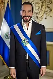 President of El Salvador Nayib Bukele wearing the presidential sash with the national flag standing behind