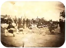 An old photograph showing a procession passing between lines of soldiers with tents in the background