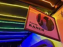Photograph of a sign with a silhouette of a man wearing a hat as well as the text "Pacos Ranch"