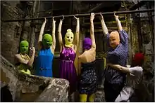 A group of 6 women wearing ski masks and multicolored clothes in a small, grubby brick space. Four of them hang by their arms from a bar, one flexes her muscles, and another grabs one of the hanging women around the waist.