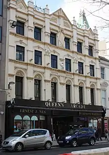 An ornate Victorian 4-storey building, painted pastel yellow, featuring muted copper spires on the roof, a small white clockface in the centre of the 2nd floor, and ground floor shopping units framed in black, either side of the arched entrance to an arcade. On the third floor is an Art-Deco-influenced monogram with the letters "A.R.".