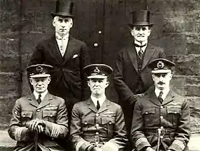 Portrait of five men, three seated wearing dark military uniforms with peaked caps, and two standing behind wearing formal suits with top hats