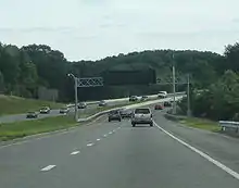 Ground-level view of a four-lane divided highway with a wide grassy median separating the opposing lanes of traffic; in the distance the road makes a sharp curve. A black electronic sign without lettering is visible above the highway.