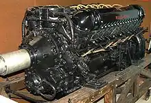A front right view of a black-painted aero engine, the words 'Rolls-Royce' appear in red. The engine has yellow electrical wiring and is sitting on a wooden pallet.
