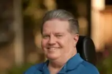 Ralph Braun, an older white man with gray hair smoothed back, wearing a blue polo, sitting in his wheelchair and smiling.