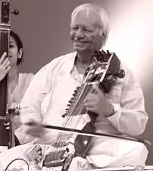 An old man sits cross-legged on a platform and plays a bowed instrument.