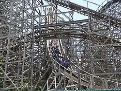 A blue train of the Rampage roller coaster is depicted reaching the bottom of the first drop to enter the slight left turn. The train is surrounded by winding wooden track and support structure, with several parts above the drop and in the background.