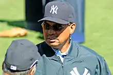 An African American male in his sixties wearing a cap and jacket, both with a logo of an overlapping "N" and two overlapping "Y"s, and sunglasses, stands on a baseball field.