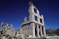 Ruins of a three-story masonry building rise into a cloudless, dark blue sky. The building is roofless, and large sections of its walls are missing. Masonry rubble lies about the building, which has openings for doors and windows but no glass or wood. Another ruin is nearby, and barren hills are visible in the distance.