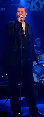 Richard Müller wearing dark clothing, standing on a dimly lit stage, appearing to sing into a microphone